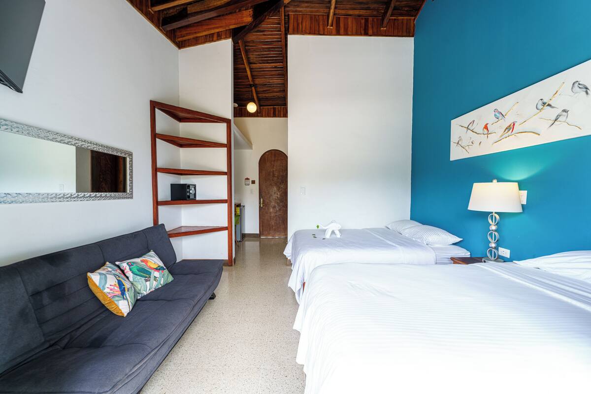 A hotel room with a sofa, two beds, a mirror, a wooden shelf, and a wall art piece.