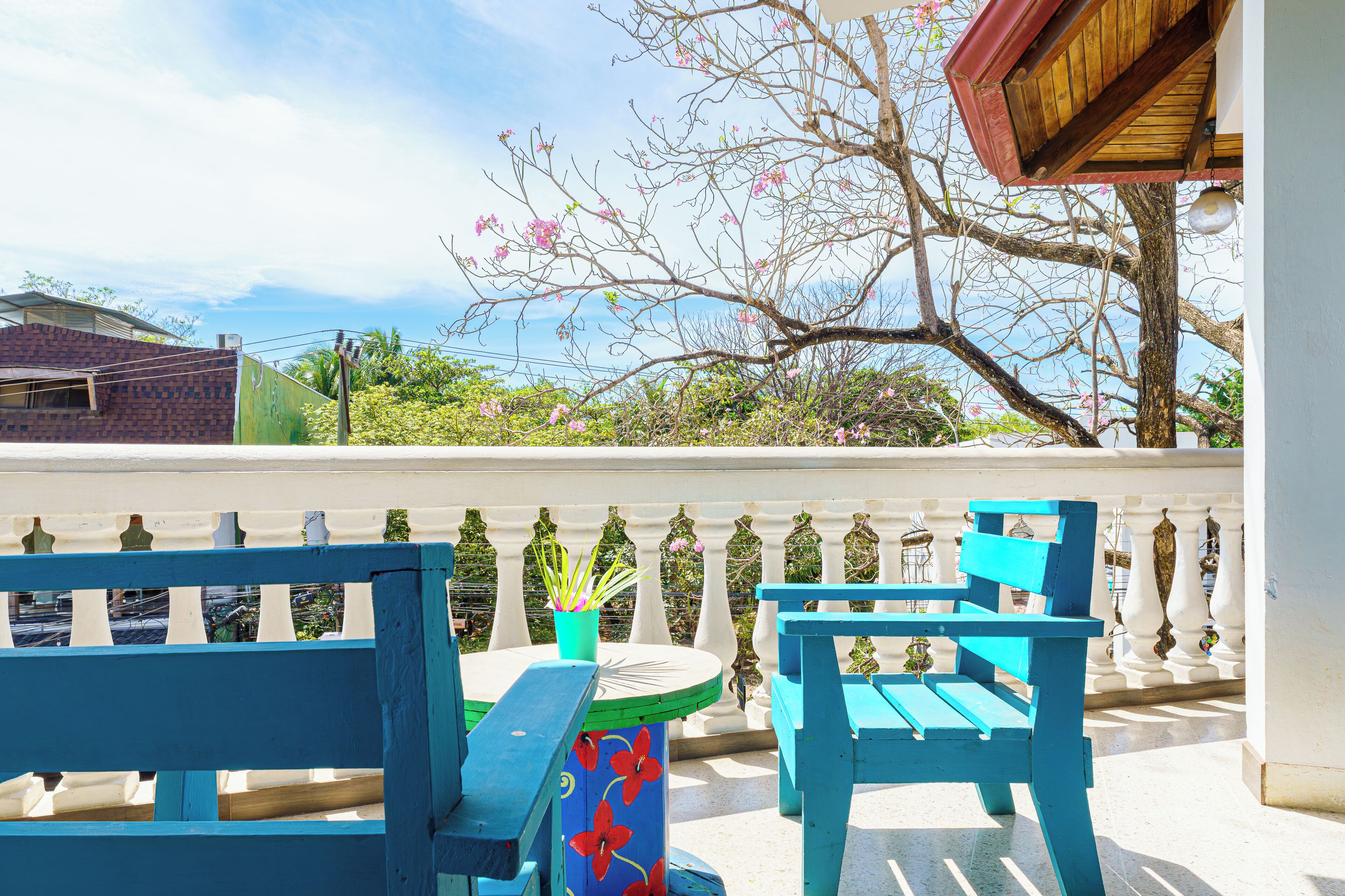A balcony with blue chairs and a table, overlooking a garden with flowering trees.