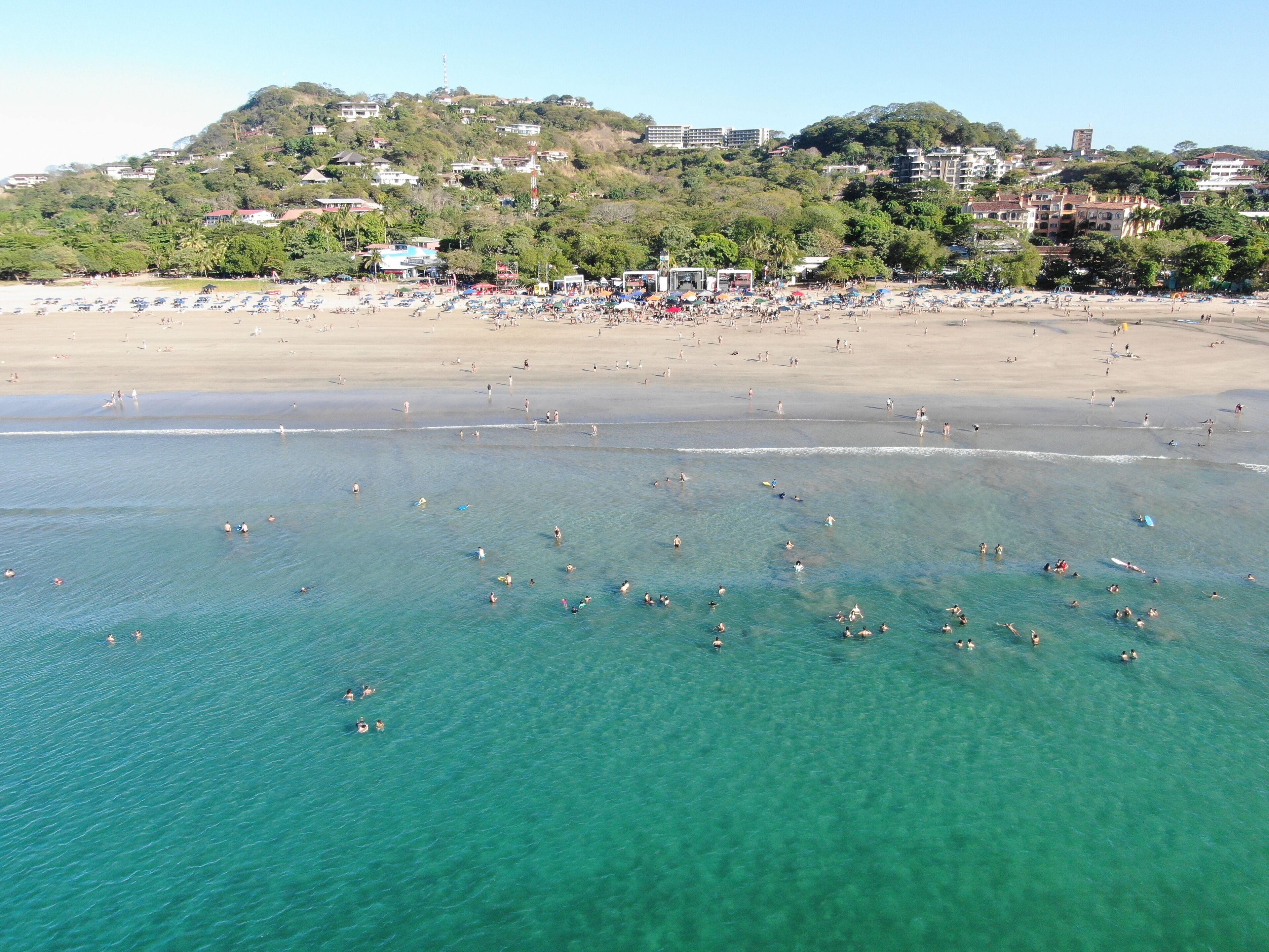 A beach with clear turquoise water, a sandy shore, and buildings in the background.