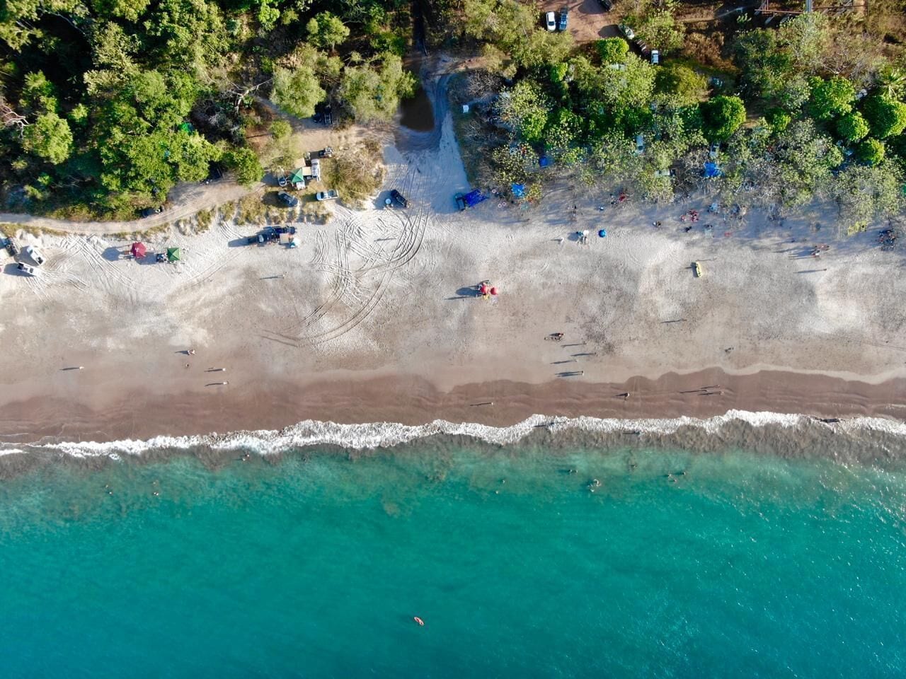 An aerial view of a beach with clear turquoise waters, a sandy shore, and scattered beachgoers.