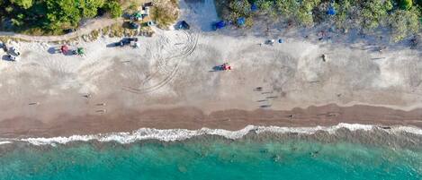 An aerial view of a beach with clear turquoise waters, a sandy shore, and scattered beachgoers.