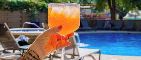 A hand holding a cocktail glass with ice cubes, a pool, and lounge chairs in the background.