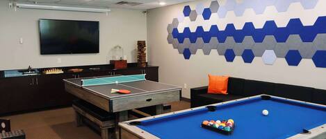A pool table, ping pong table, and a large flat-screen TV in a room with a hexagonal wall design.