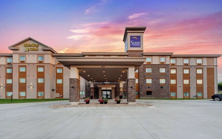 A MainStay Suites hotel with a modern entrance, a parking lot, and a clear sky.