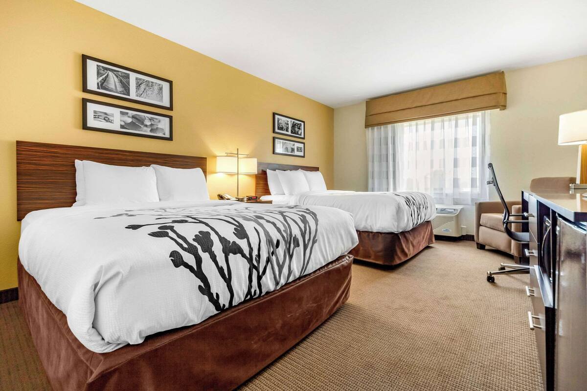 A hotel room with two beds, a desk, a chair, and a lamp.