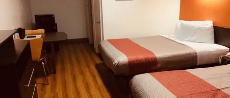 A hotel room with a wooden floor, a bed with a red and grey bedspread, a desk with a chair, and a wall-mounted light fixture.
