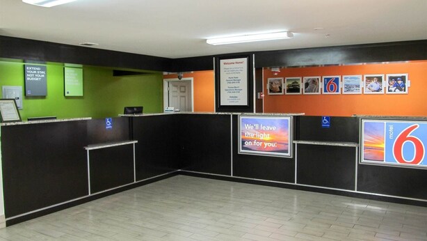 A reception area with a black counter, green and orange walls, and a sign that says "We'll leave the light on for you." - Motel 6 Augusta, GA - Fort Gordon (Augusta)