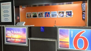 Zona de recepción de un hotel con un letrero que indica el nombre 'motel 6' y un letrero de accesibilidad para personas con discapacidad. Hay cuadros enmarcados en la pared y una pantalla digital que muestra una imagen de atardecer y texto.