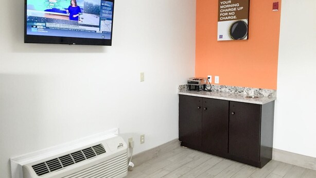 Una habitación con un televisor en la pared mostrando un canal de noticias, una pequeña cocina con microondas y un aire acondicionado instalado en la pared.