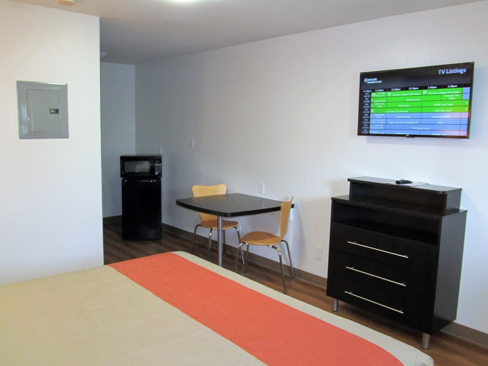 A hotel room with a bed, a TV mounted on the wall displaying TV listings, a small table with two chairs, and a kitchenette area with a microwave.