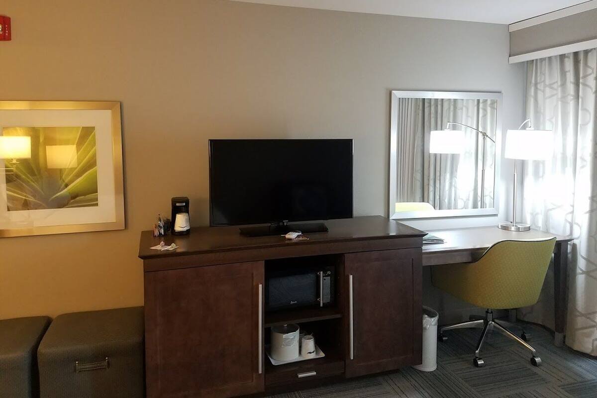 A hotel room with a flat-screen TV, a desk with a chair, a framed picture on the wall, and a window with curtains.
