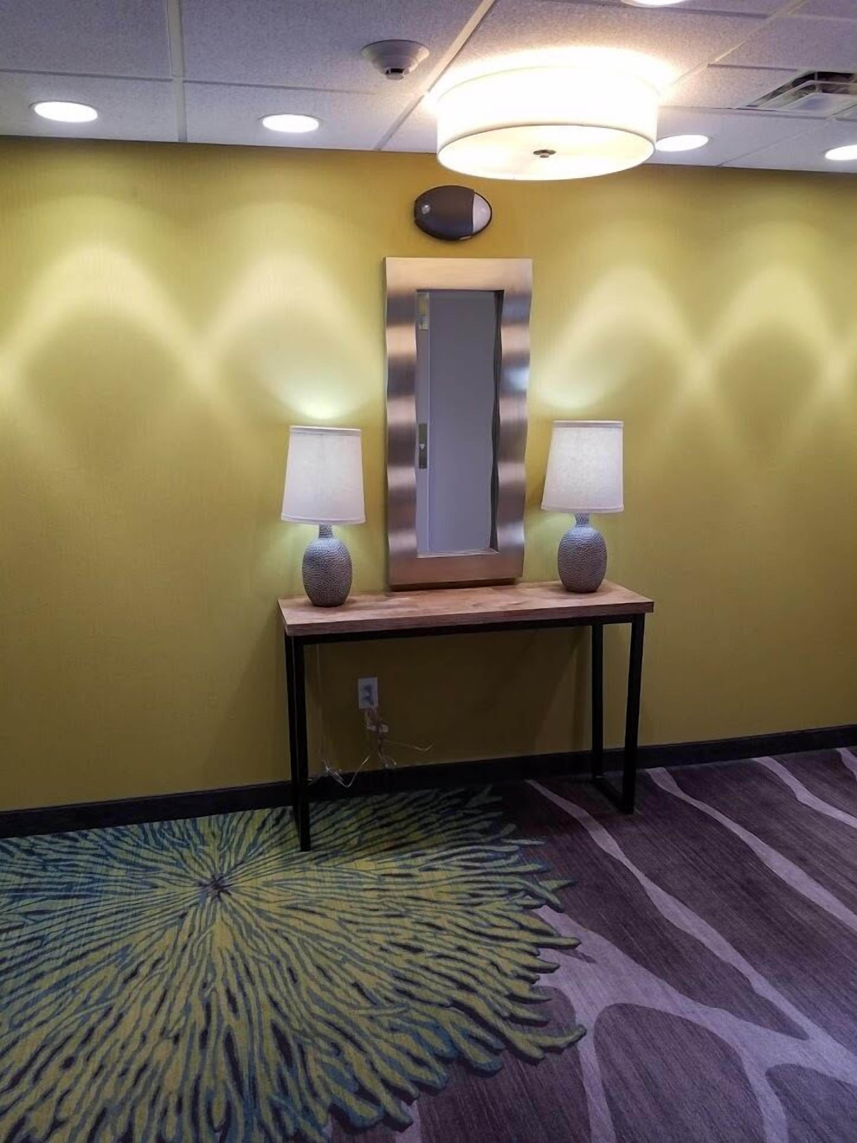 Habitación de hotel con un espejo sobre una consola de madera, dos lámparas a cada lado y una alfombra estampada.