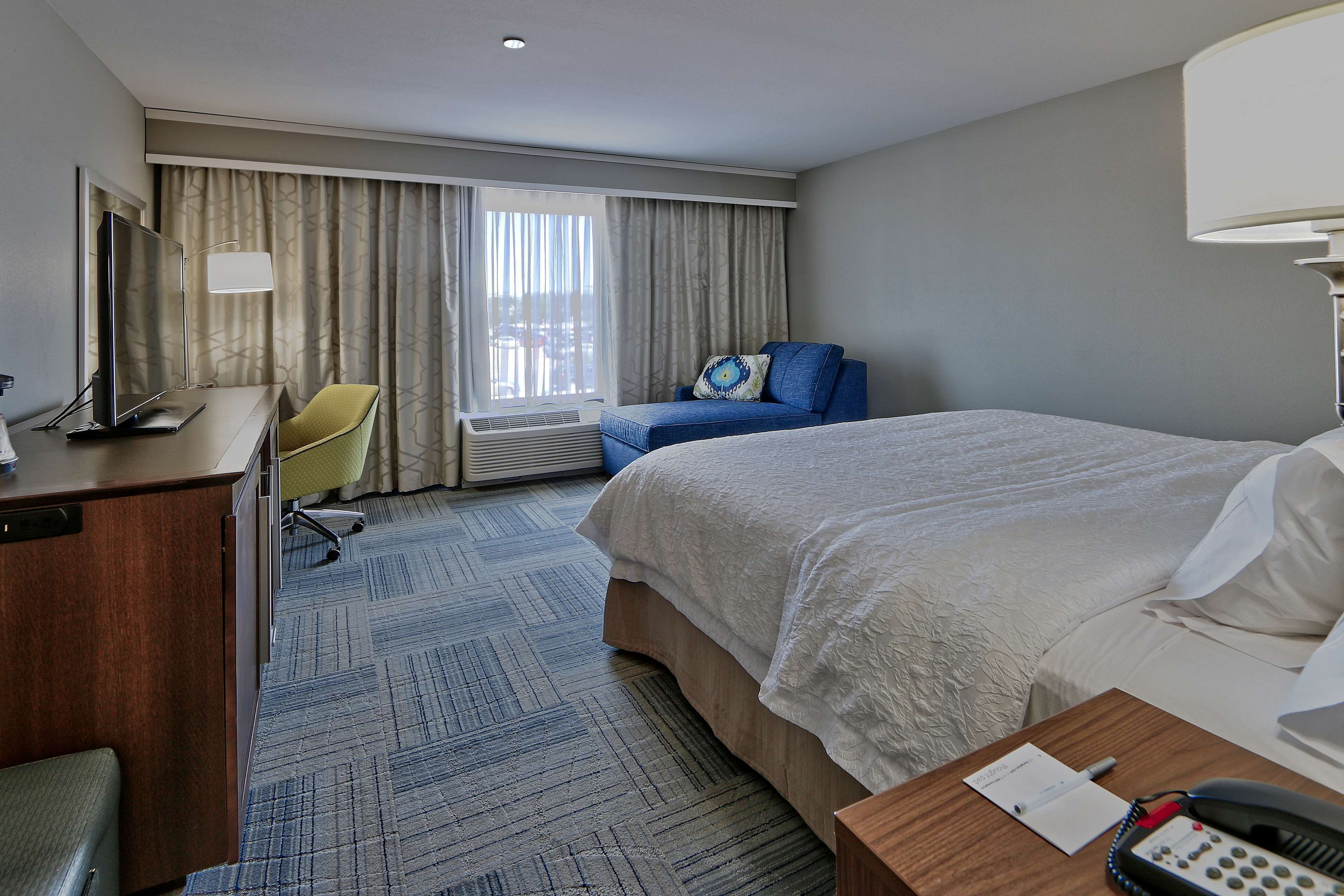 A hotel room with a bed, a desk with a computer, a chair, a lamp, and a window with curtains.