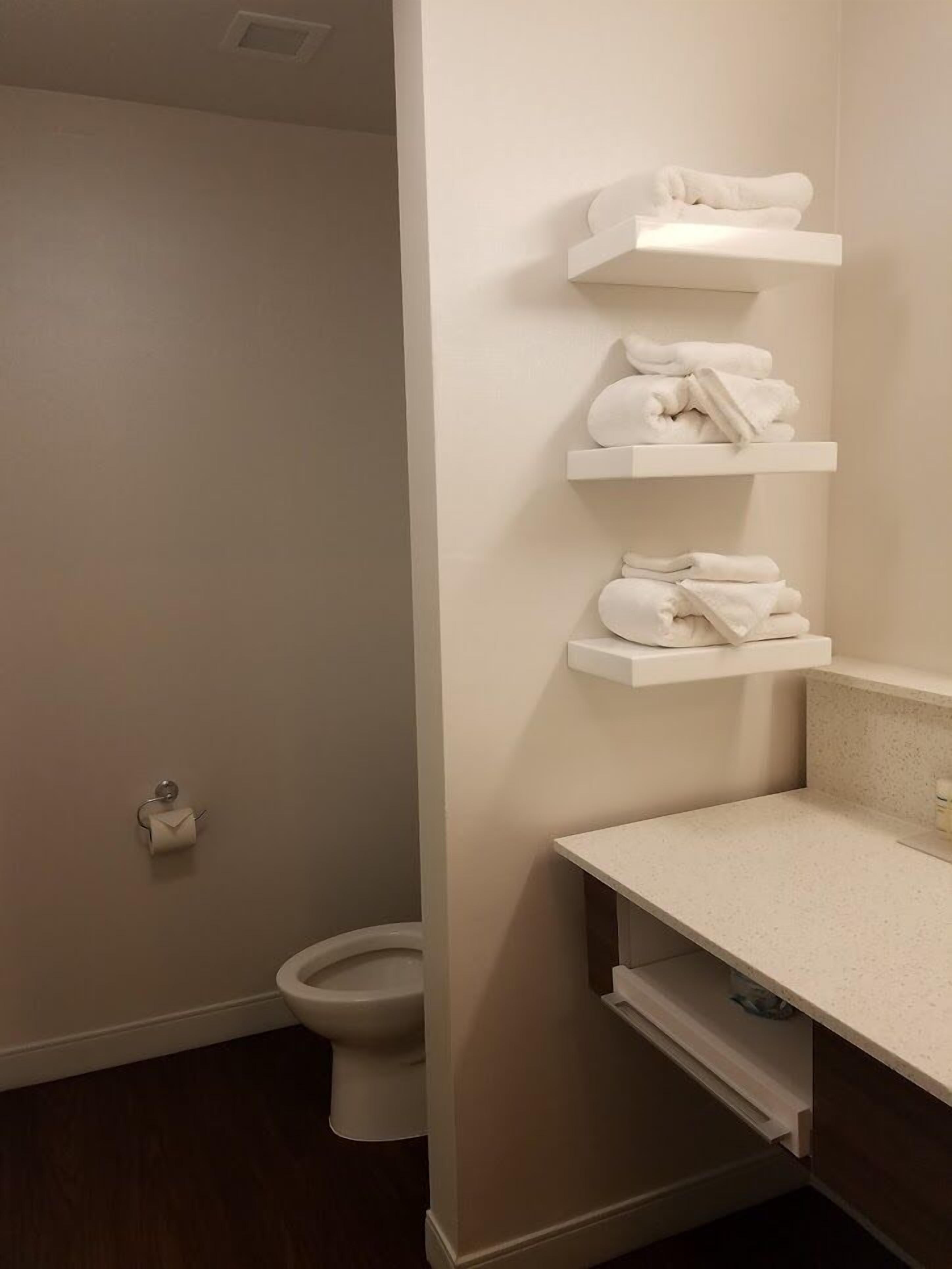 Une salle de bain aux murs blancs, avec des toilettes et trois étagères blanches pour les serviettes.