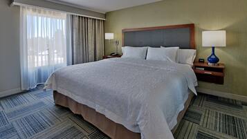 A hotel room with a large bed, two bedside tables, a nightstand, a lamp, and a view of the outside through sheer curtains.