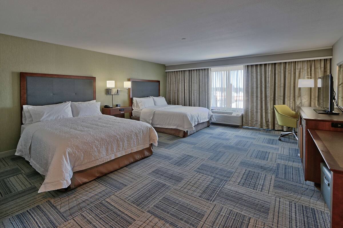A hotel room with two beds, a desk, a chair, and a large window with curtains.