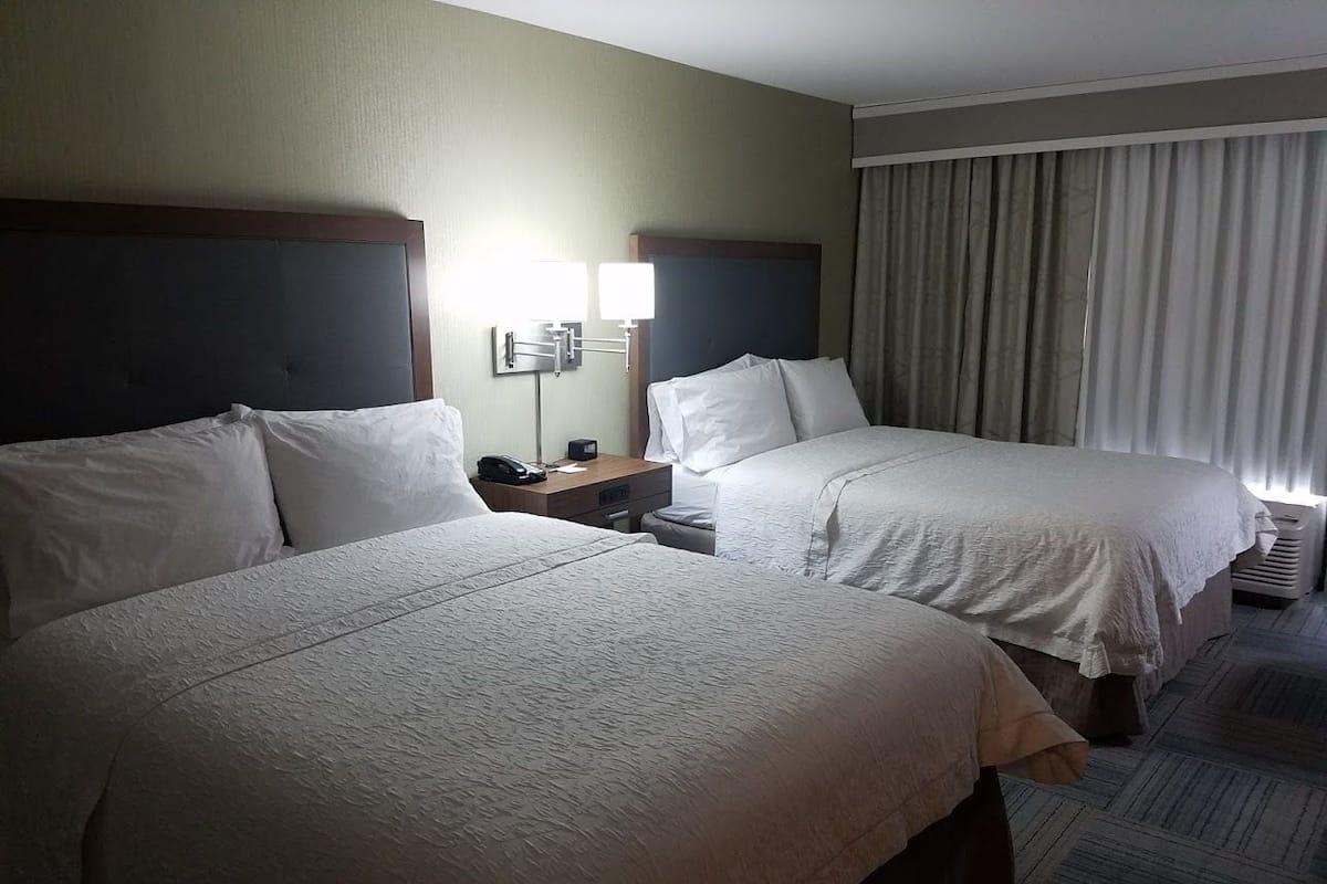 A hotel room with two beds, a nightstand, a lamp, and a window with curtains.