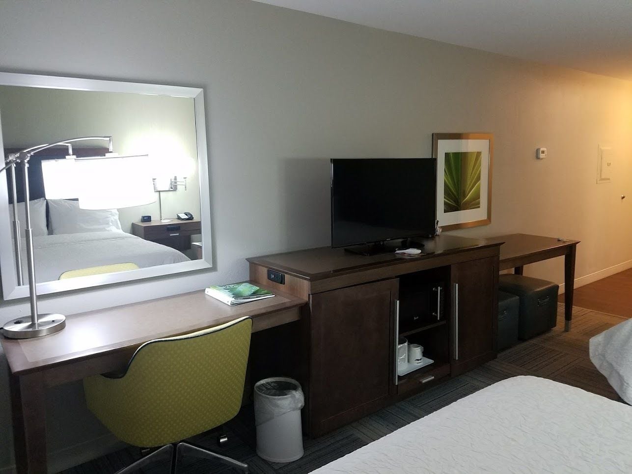 Une chambre d’hôtel comprenant un lit, un bureau avec une chaise, une télévision, un miroir, une poubelle et un tableau encadré au mur.