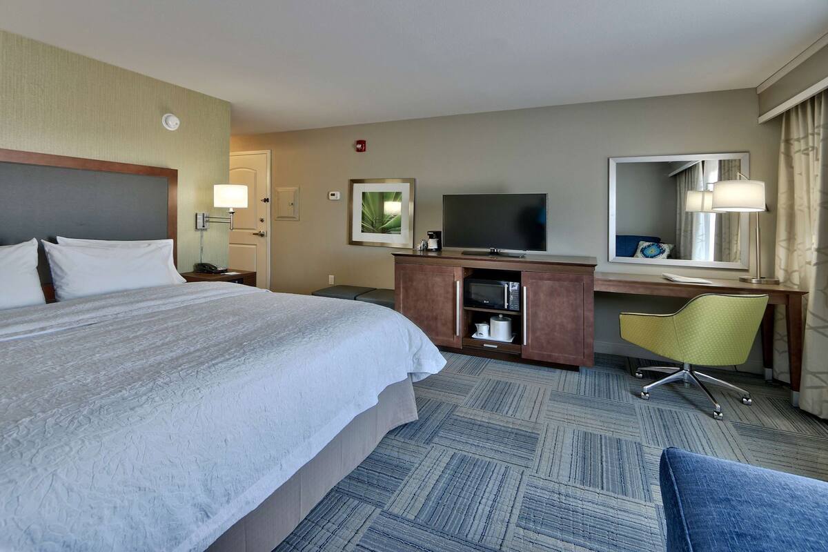 A hotel room with a large bed, a desk with a computer, a television, a chair, and a small kitchenette.
