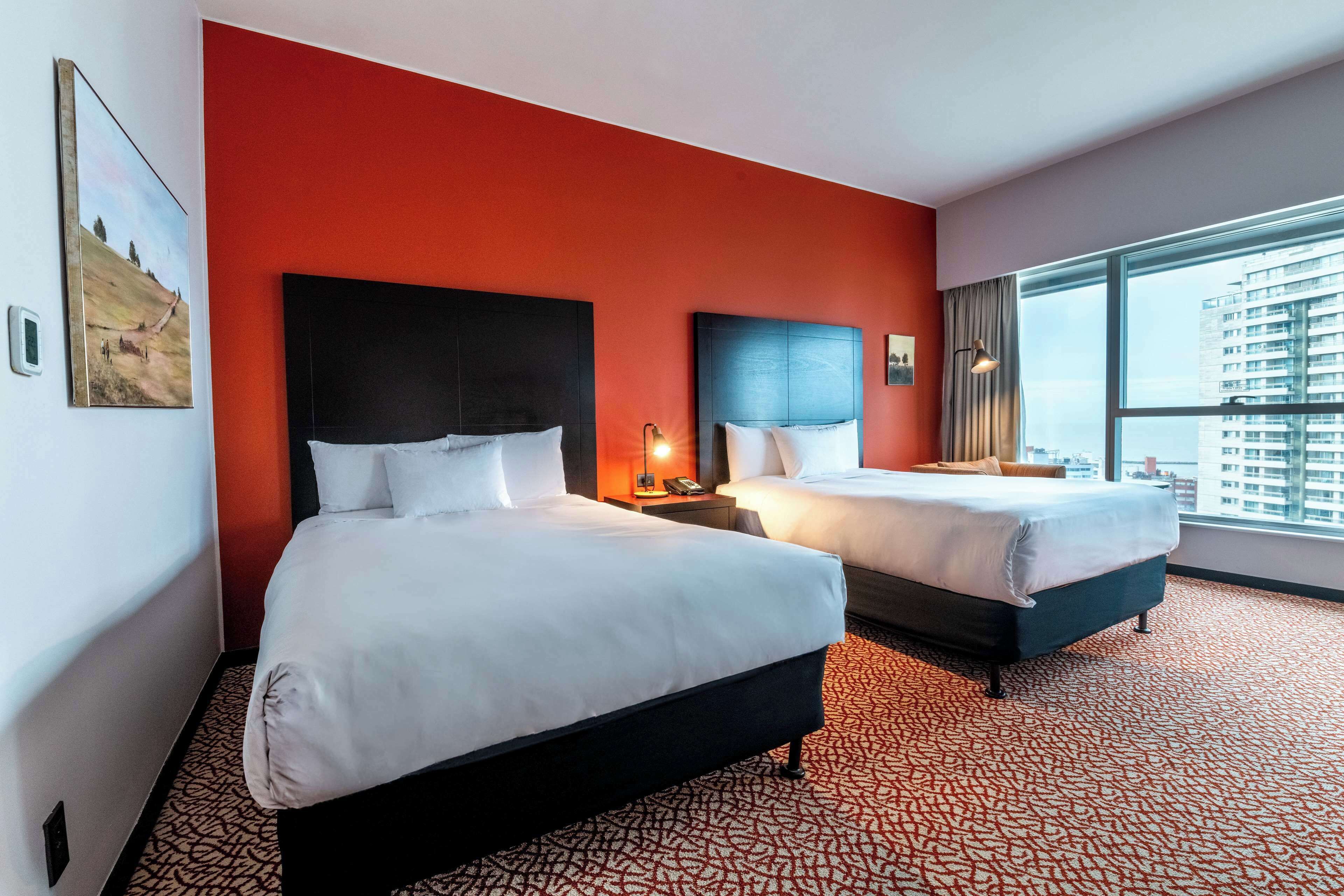 Habitación de hotel con dos camas, un muro decorativo rojo y un ventanal con vistas a la ciudad.