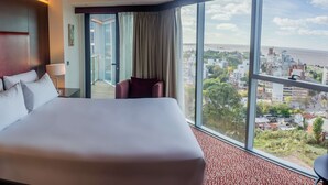 Une chambre d’hôtel avec un grand lit, un bureau avec une lampe, un téléphone et une vue sur la ville et la mer.