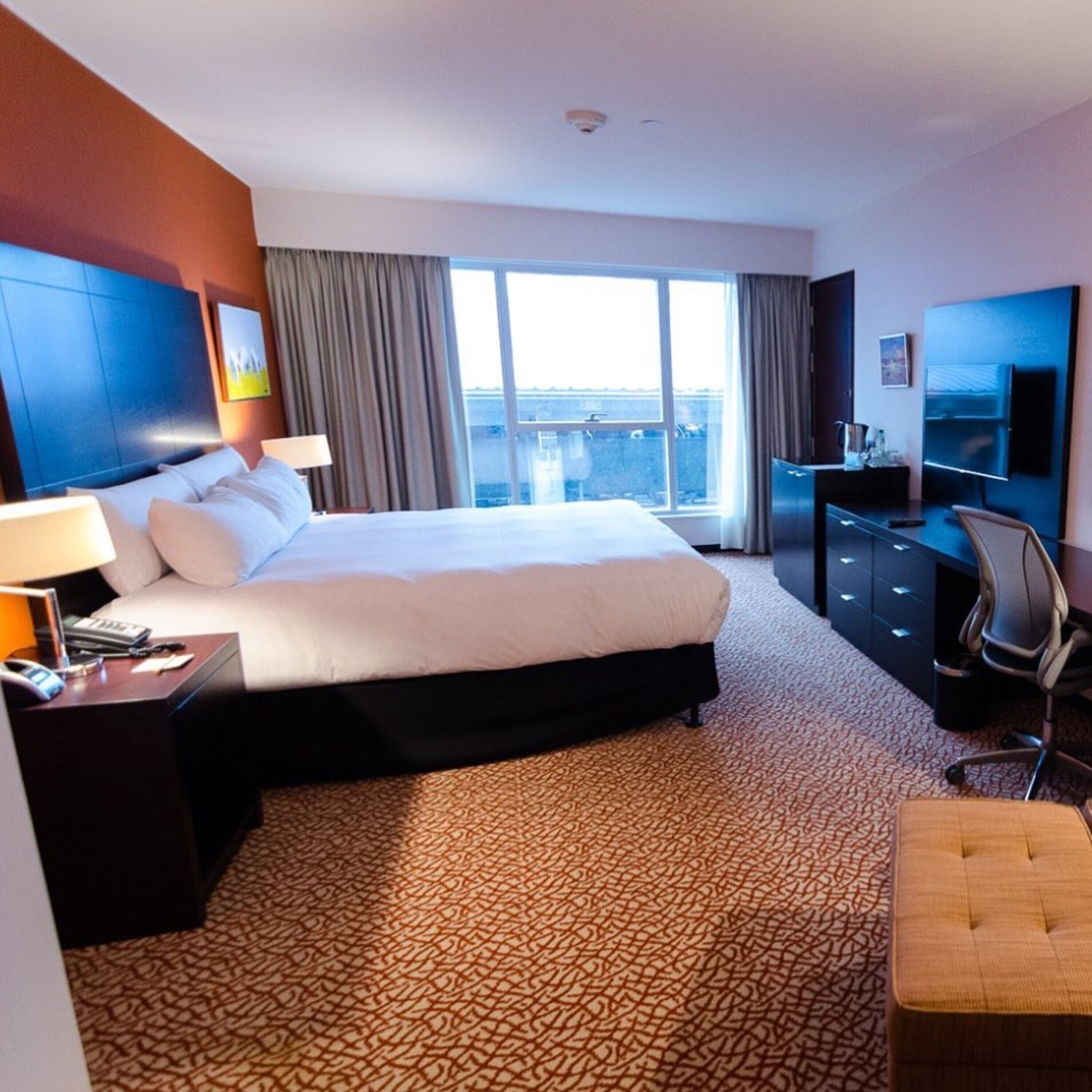 A hotel room with a large bed, a desk with a lamp, a TV, and a view of the city through the window.