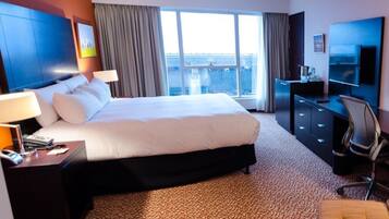 A hotel room with a large bed, a desk with a lamp, a TV, and a view of the city through the window.
