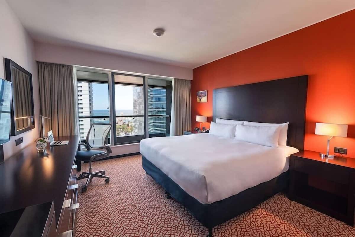A hotel room with a large bed, a desk, a chair, a TV, and a view of the city.