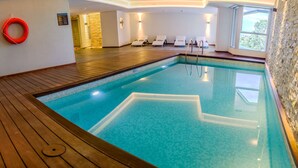 Une piscine intérieure avec un sol en bois, une eau d’un bleu cristallin et des chaises longues blanches.