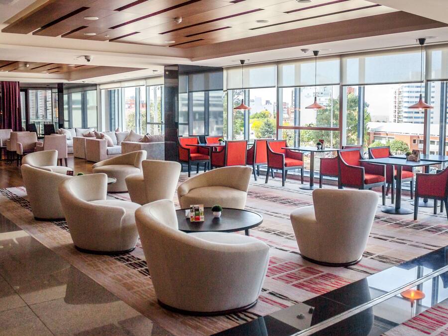 Um saguão de hotel moderno com diversas áreas de estar, incluindo sofás e poltronas, e uma área de refeições com mesas e cadeiras. Grandes janelas oferecem vista para a paisagem urbana.