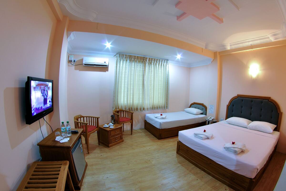 A hotel room with two single beds, a TV, a desk with a chair, a small table, and an air conditioning unit.
