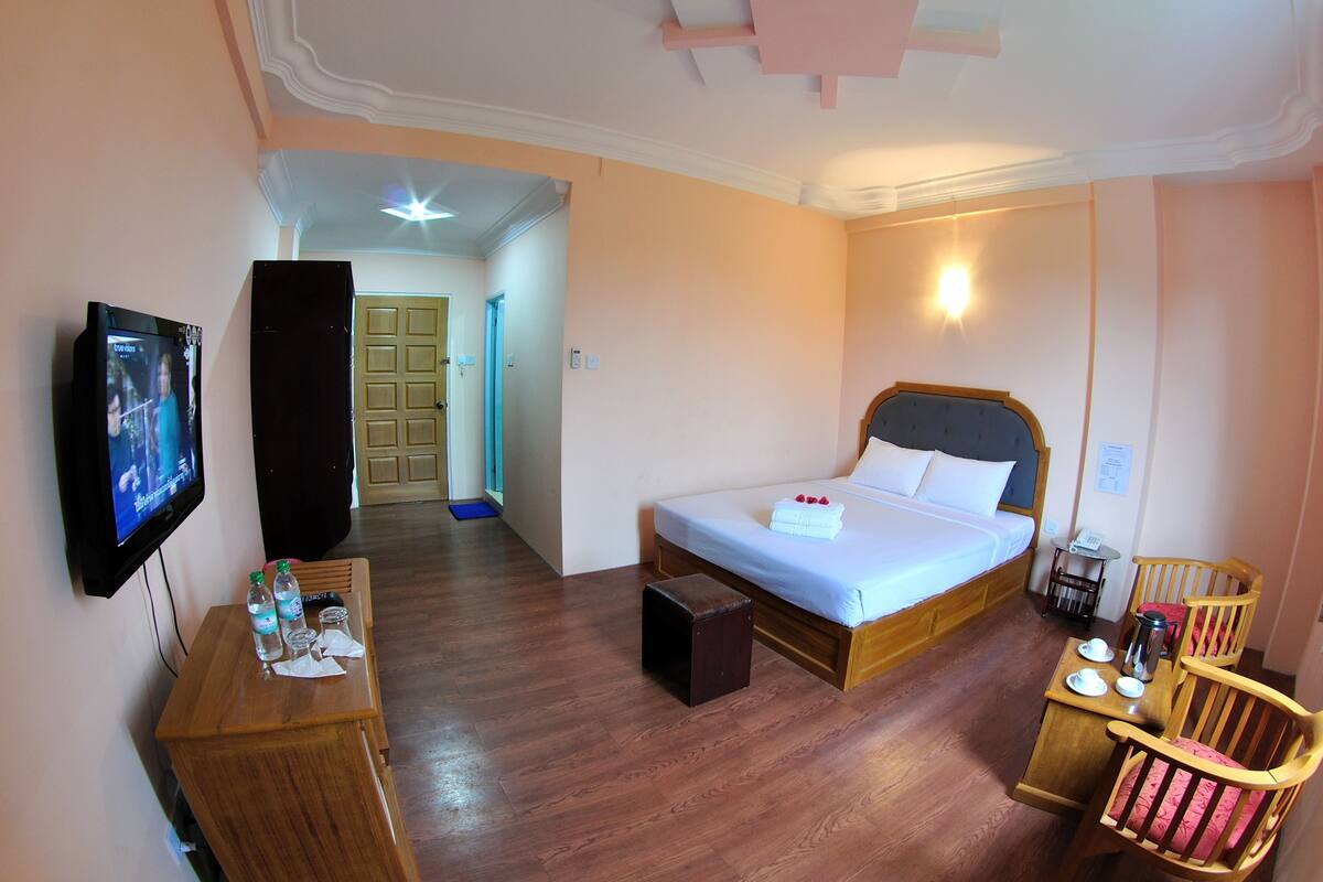 A hotel room with a bed, bedside table, TV, and a small table with cups and a thermos.