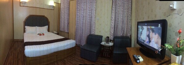 Quarto de hotel com uma cama, duas poltronas, uma mesinha com um comando remoto e uma televisão exibindo um programa.