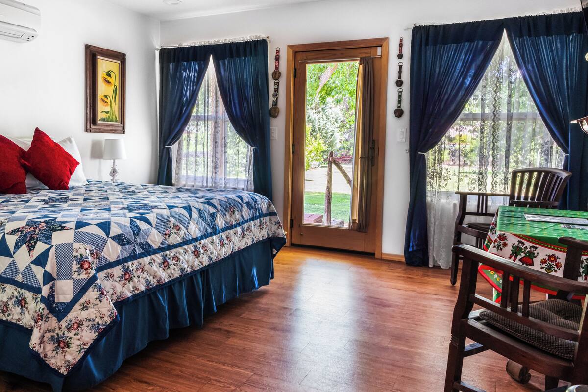 Une chambre avec un plancher en bois, un lit recouvert d’une couette bleue et blanche, des oreillers rouges, une chaise en bois et une porte donnant sur un balcon.