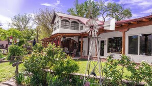 Una casa de dos pisos con un molino, un porche con mobiliario exterior y un jardín con diversas plantas y flores.