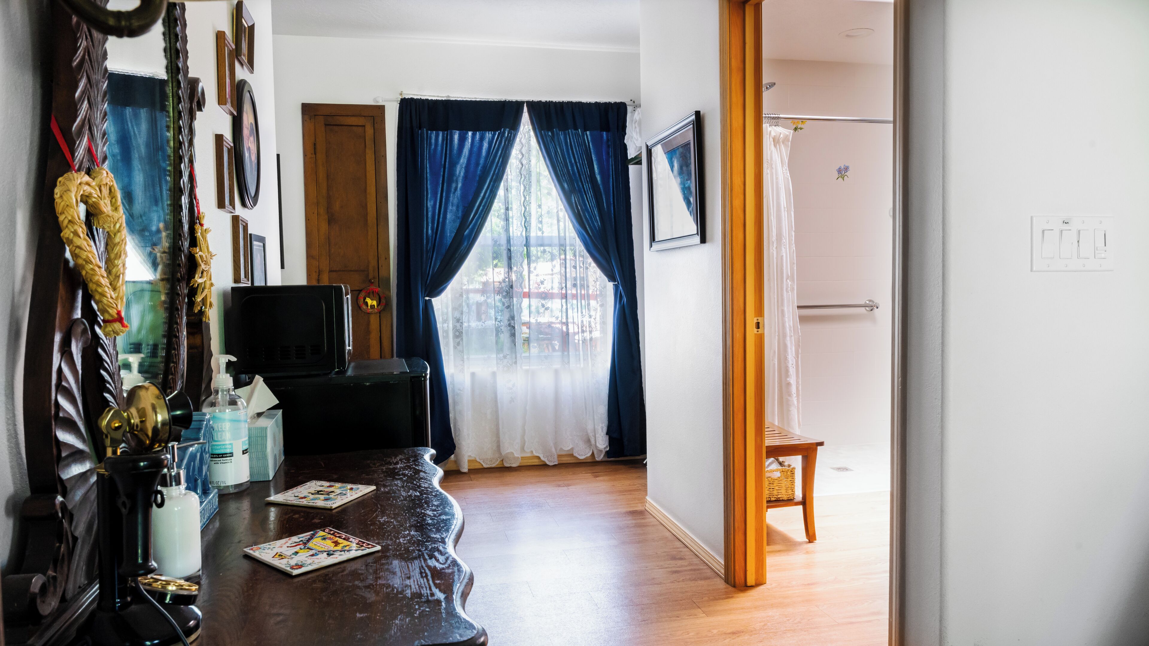 A living room with a wooden floor, a television, and a wooden table with a bottle and a box. There is a door leading to a bathroom with a shower and a towel rack.