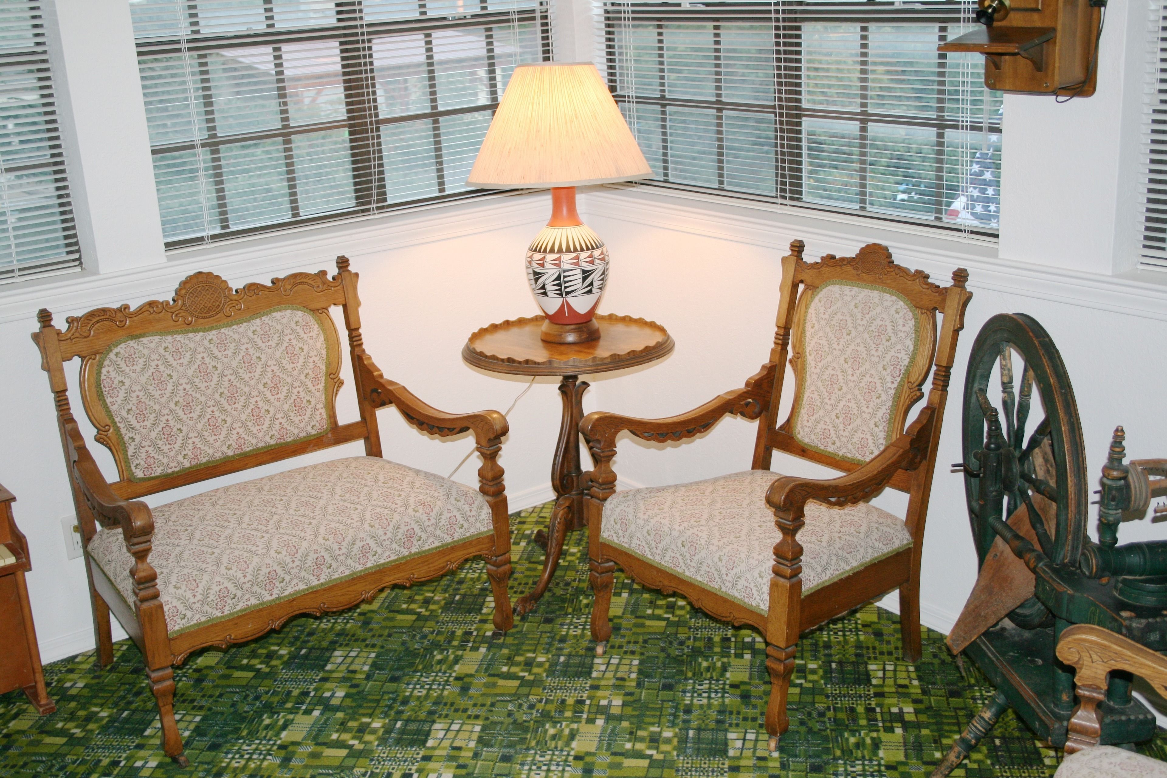 Ein Zimmer mit zwei hölzernen Stühlen, deren Sitzflächen und Rücken gepolstert sind, einem kleinen runden Tisch mit einer Lampe und einem Spinnrad.