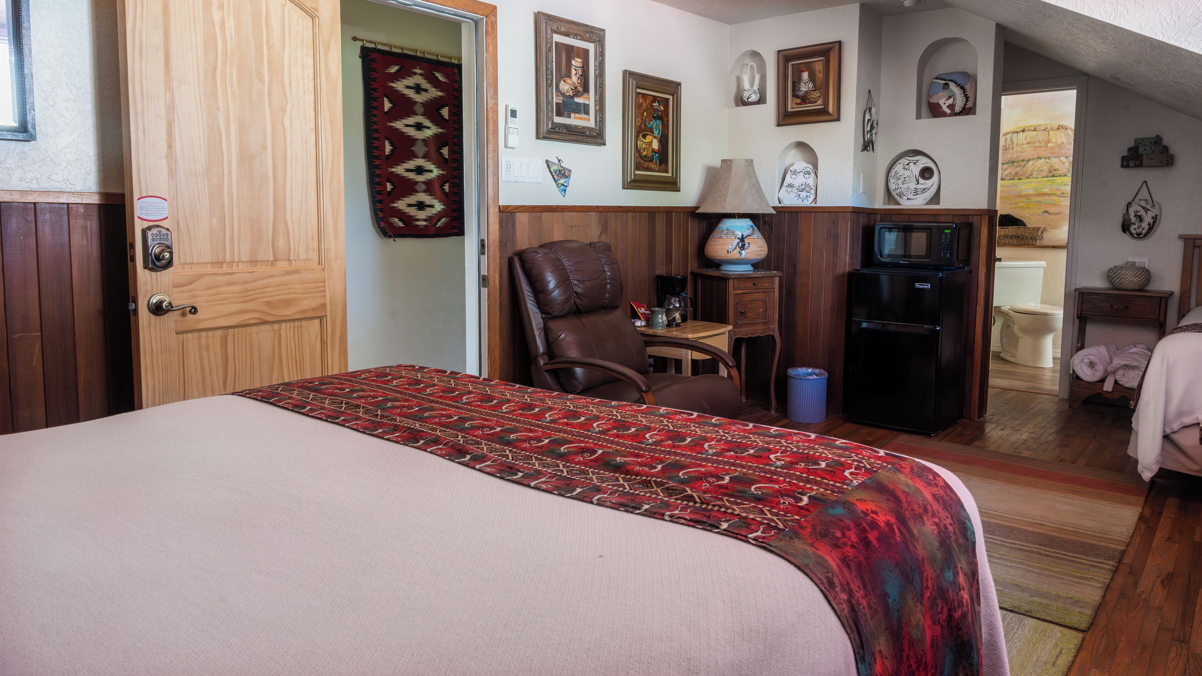 A hotel room with a bed, a desk, a chair, a refrigerator, and a bathroom visible through an open door.