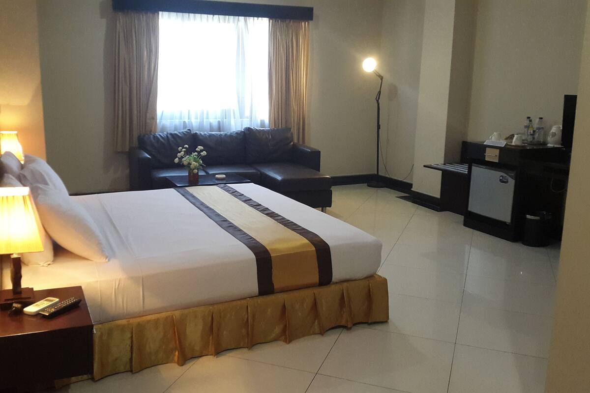 A hotel room with a bed, a desk with a lamp, a sofa, a desk with a computer, and a window with curtains.