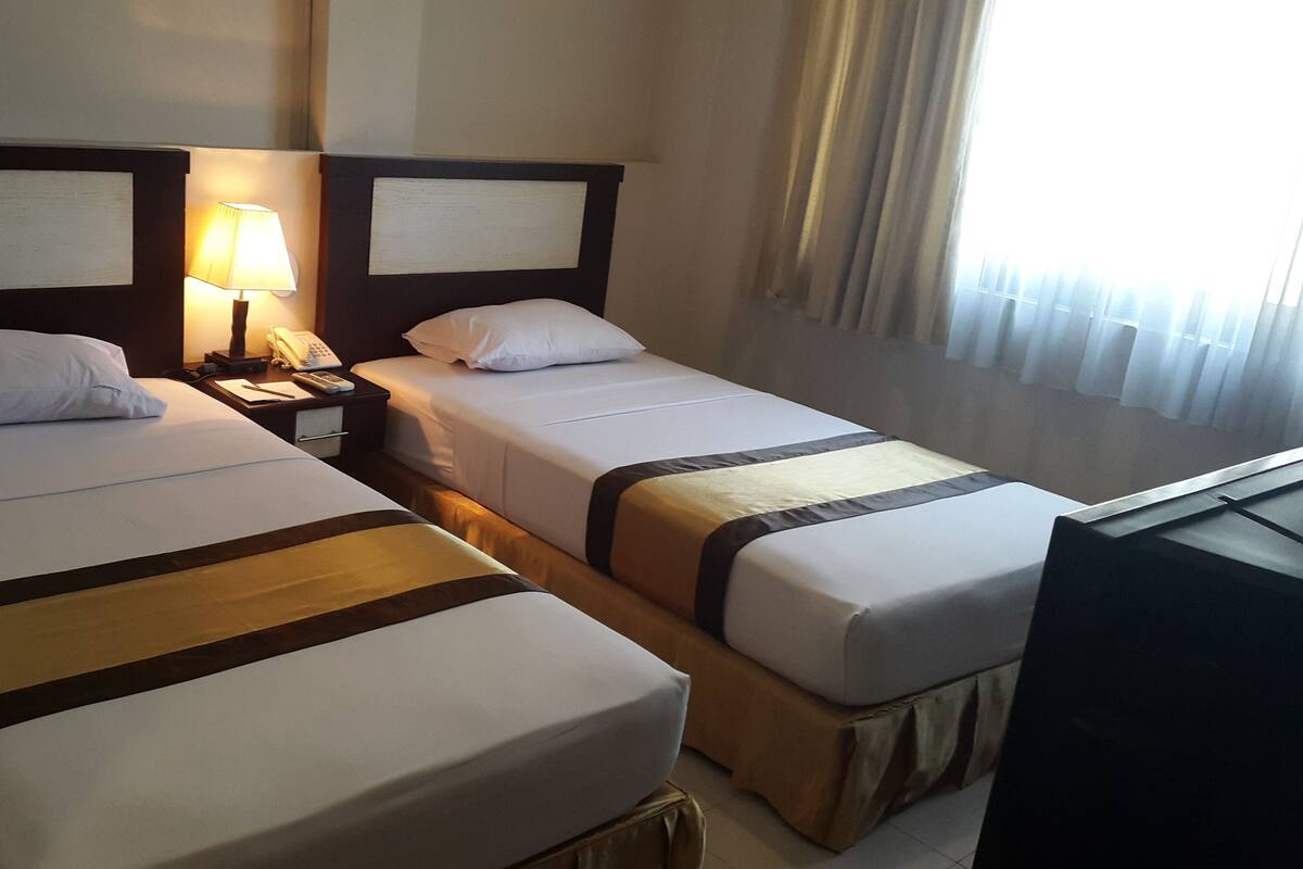 A hotel room with two single beds, a nightstand with a lamp, a telephone, and a television.