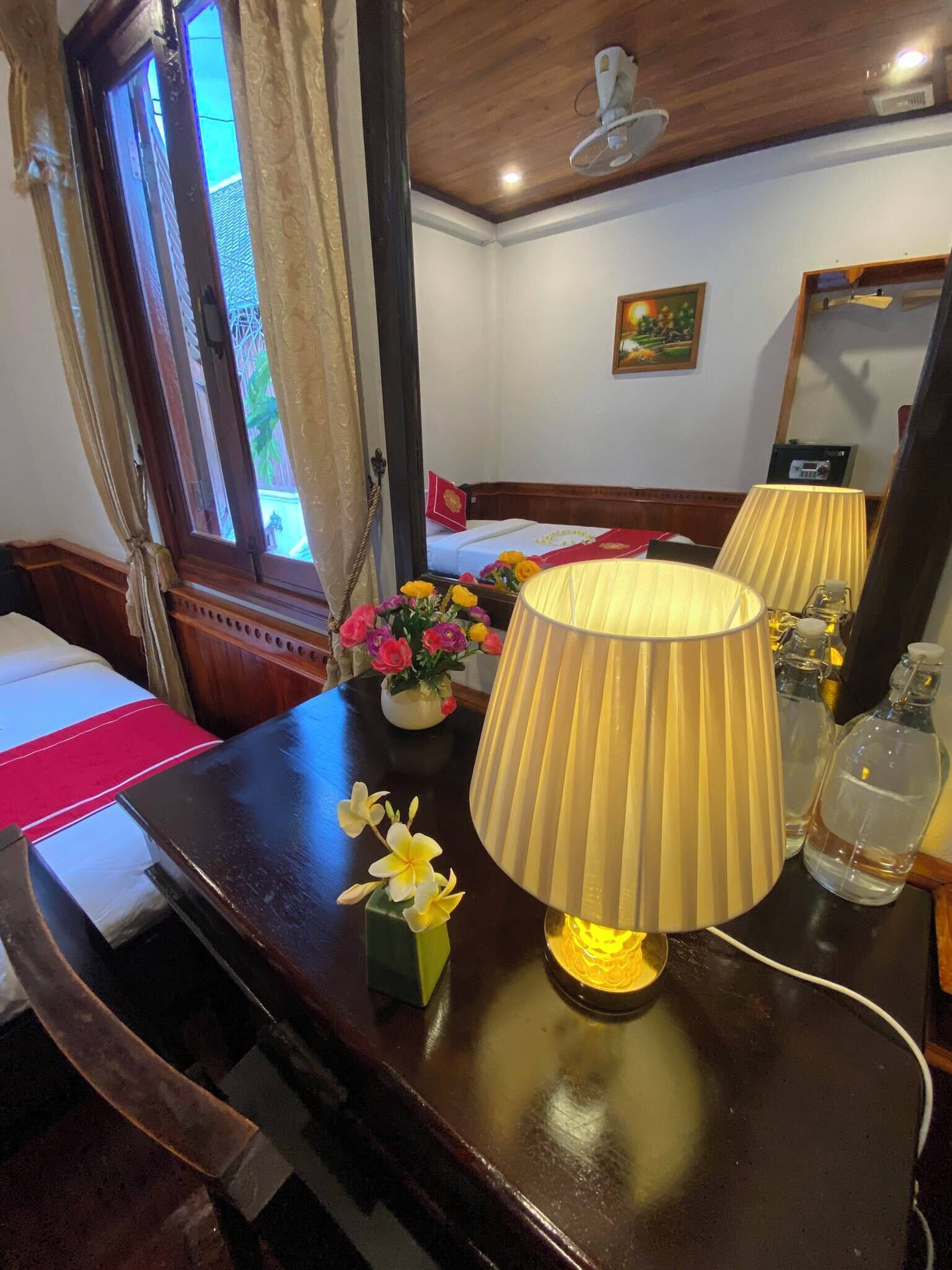 Une chambre d’hôtel avec un plafond en bois, un lit recouvert d’une courtepointe rouge et blanche, une table avec une lampe, un vase rempli de fleurs et une fenêtre avec des rideaux.