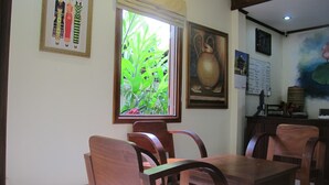 A dining area with wooden chairs and a table, a window with a view of greenery, and artwork on the walls. - Golden Lotus Place (Luang Prabang)