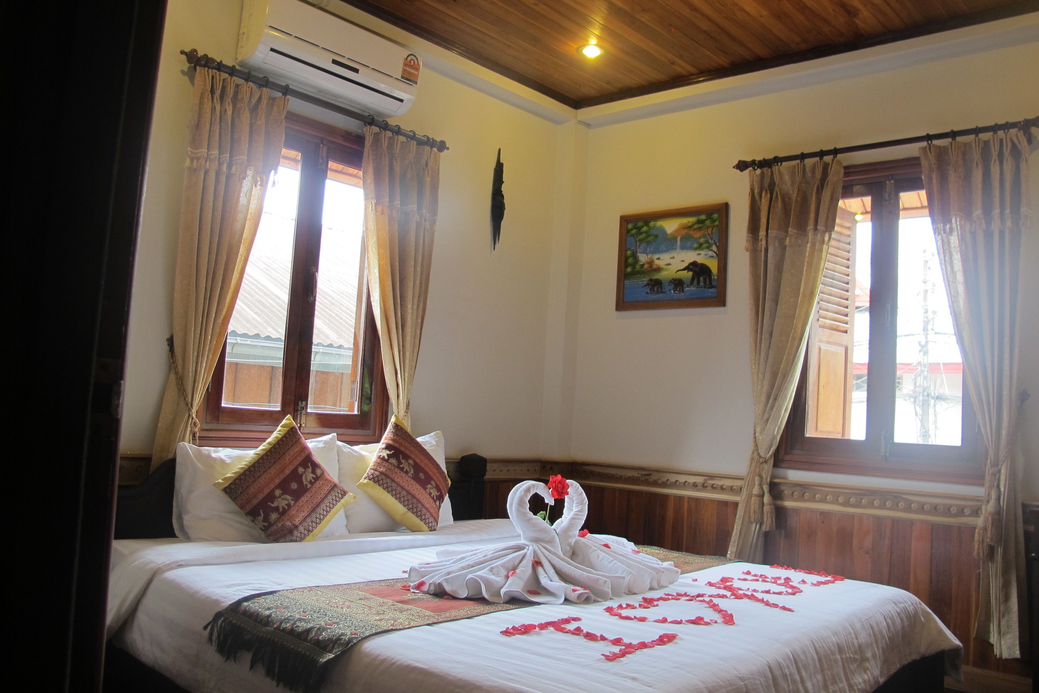 A hotel room with a bed, curtains, a painting, and an air conditioning unit.