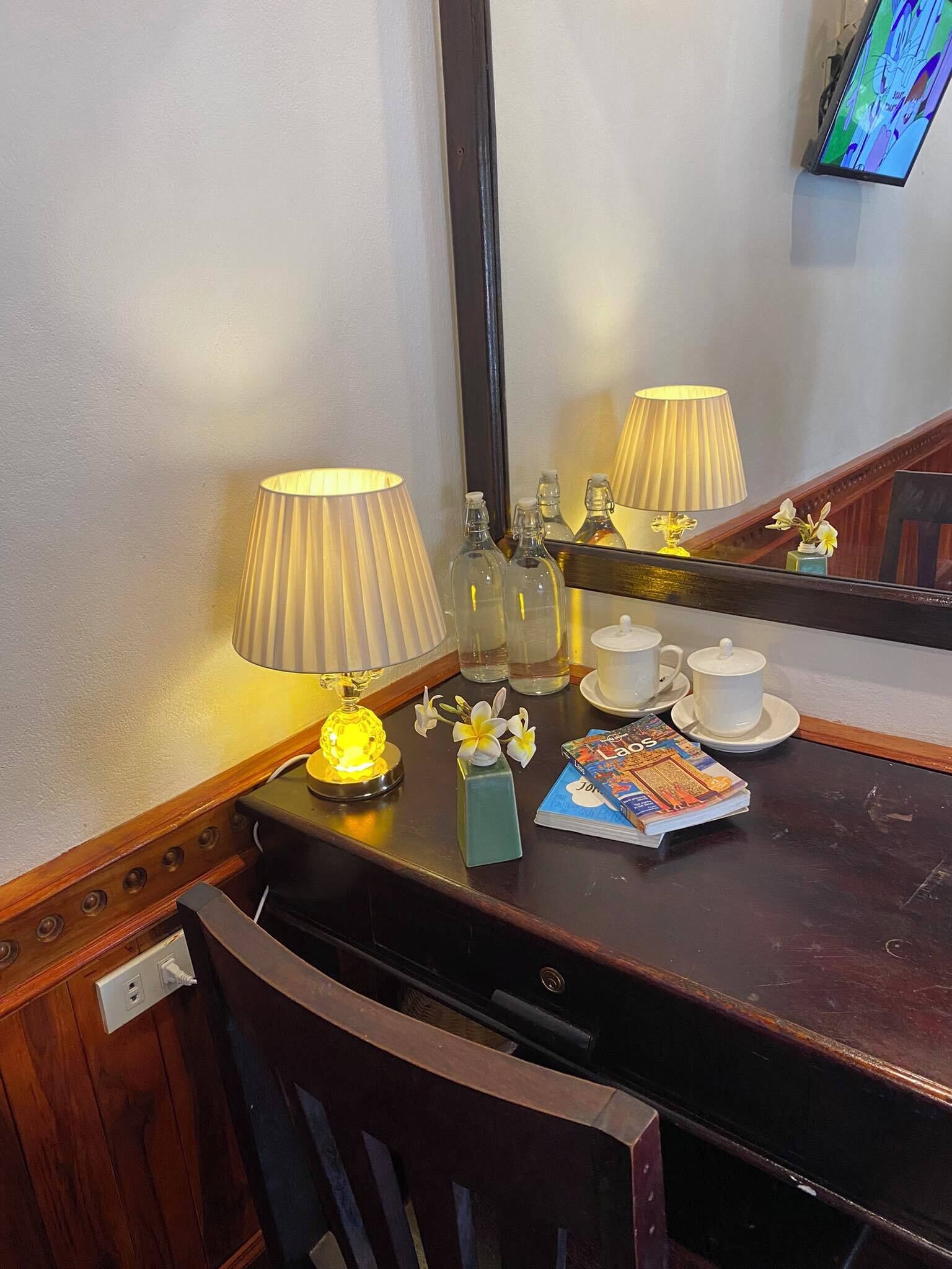 A wooden desk with a mirror, two lamps, a vase with flowers, a stack of magazines, and a tray with teacups and a teapot.