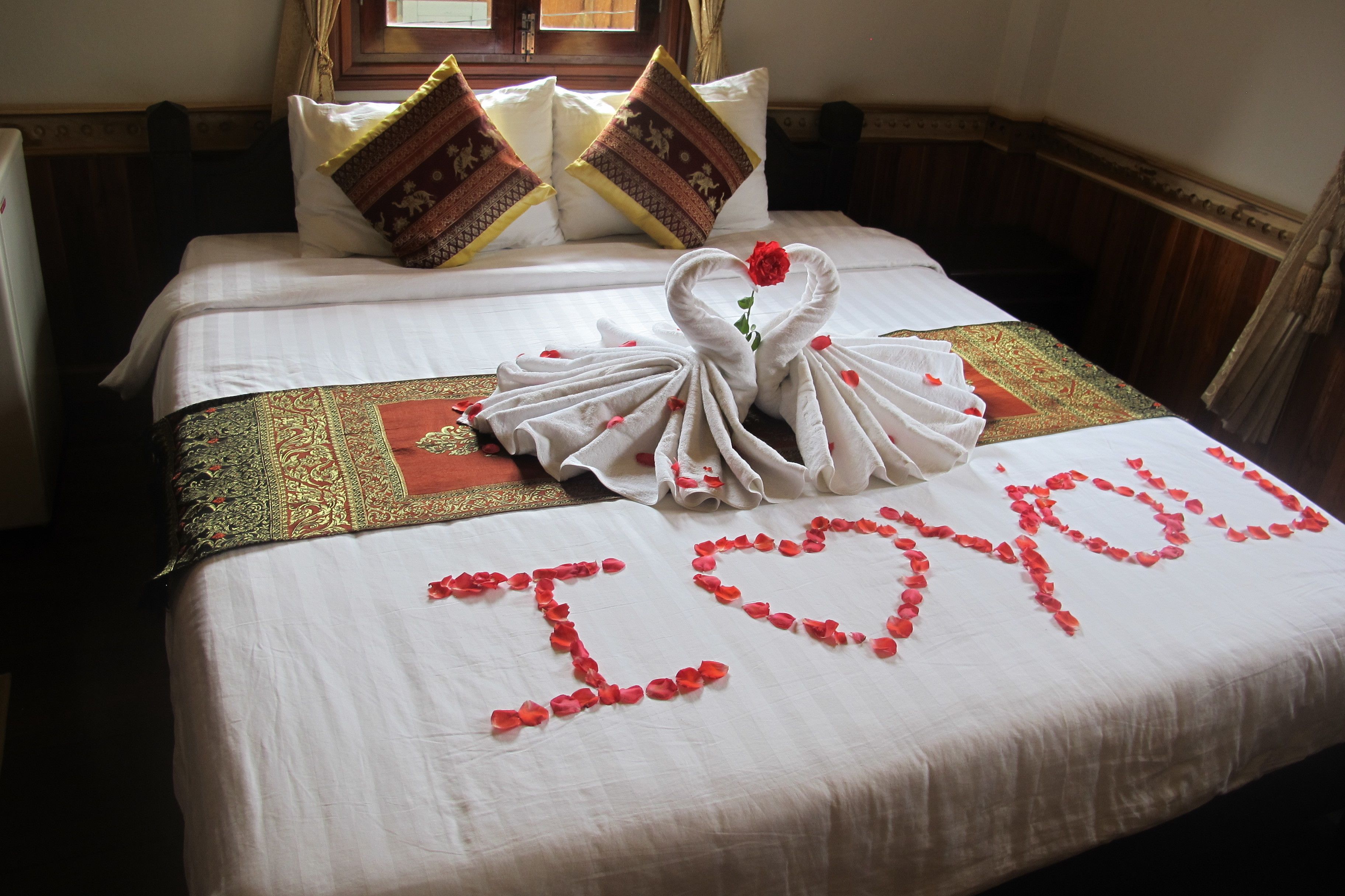A neatly made bed with a heart-shaped towel arrangement spelling "I love you" and a rose on the bed.
