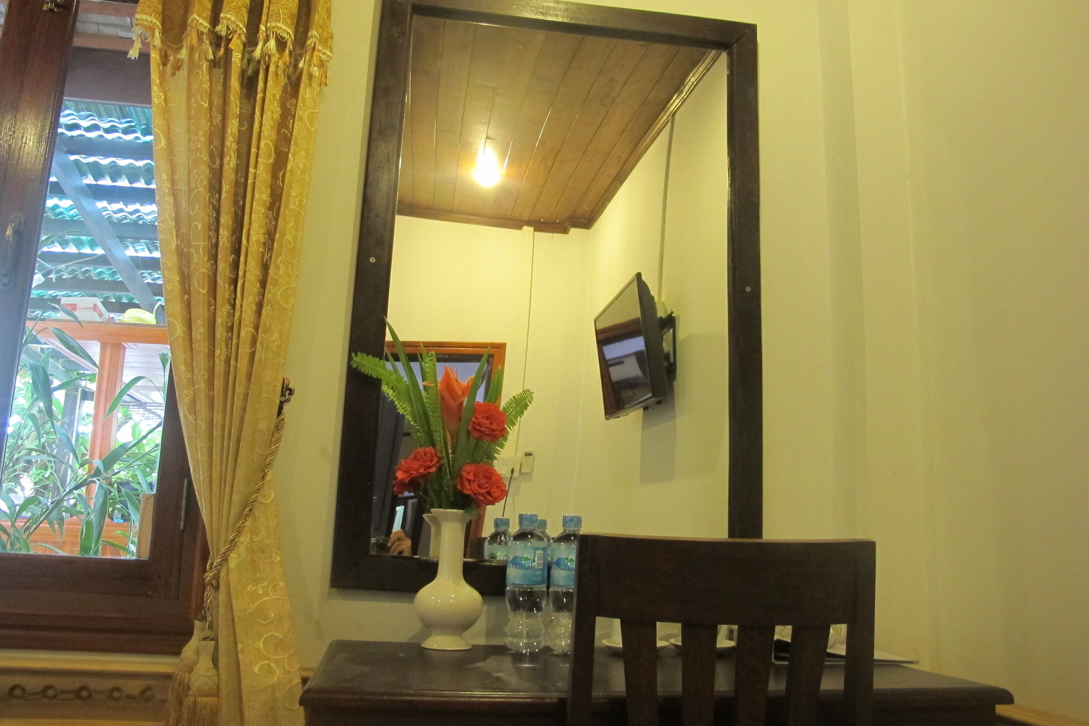 A room with a wooden ceiling, a window with curtains, a mirror, a vase with flowers, and a table with water bottles.