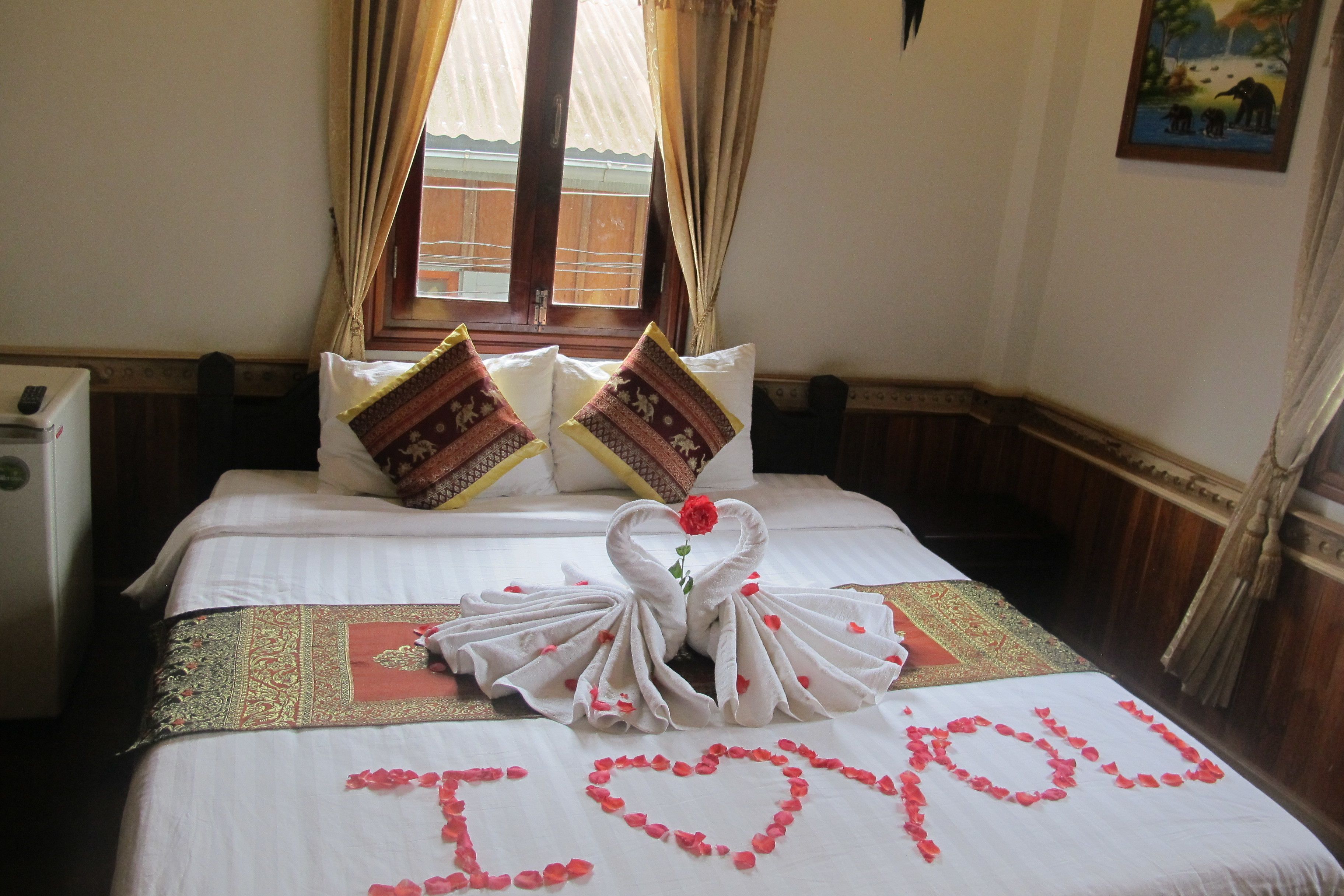 Una cama cuidadosamente tendida con una toalla con forma de cisne y pétalos de rosa que deletrean 'I love you' sobre la colcha.