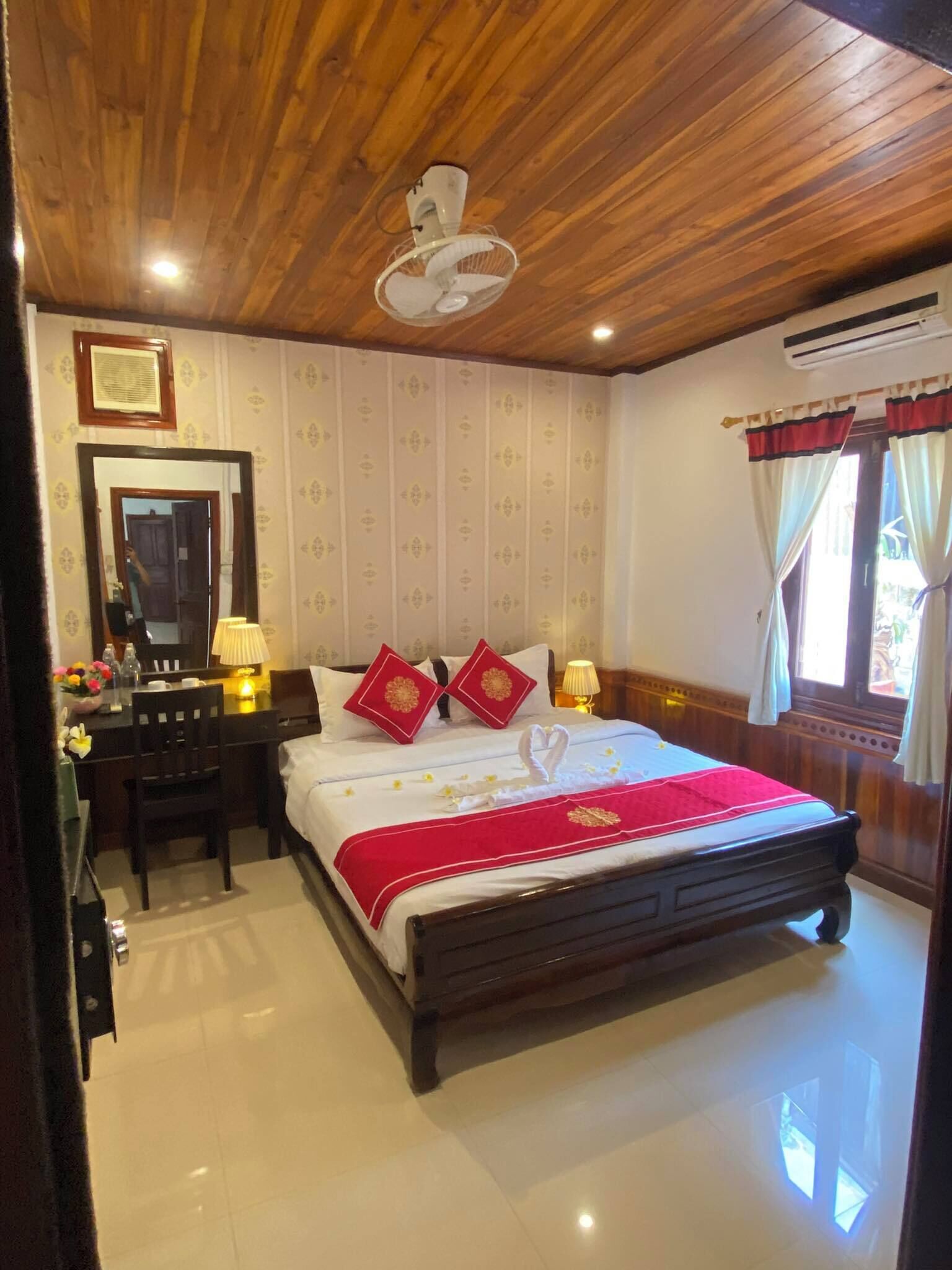A hotel room with a wooden ceiling, a bed with a red bedspread, two red pillows, a bedside table with a lamp, a mirror, and a window with curtains.
