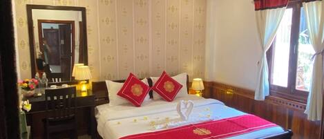 A hotel room with a wooden ceiling, a bed with a red bedspread, two red pillows, a bedside table with a lamp, a mirror, and a window with curtains.