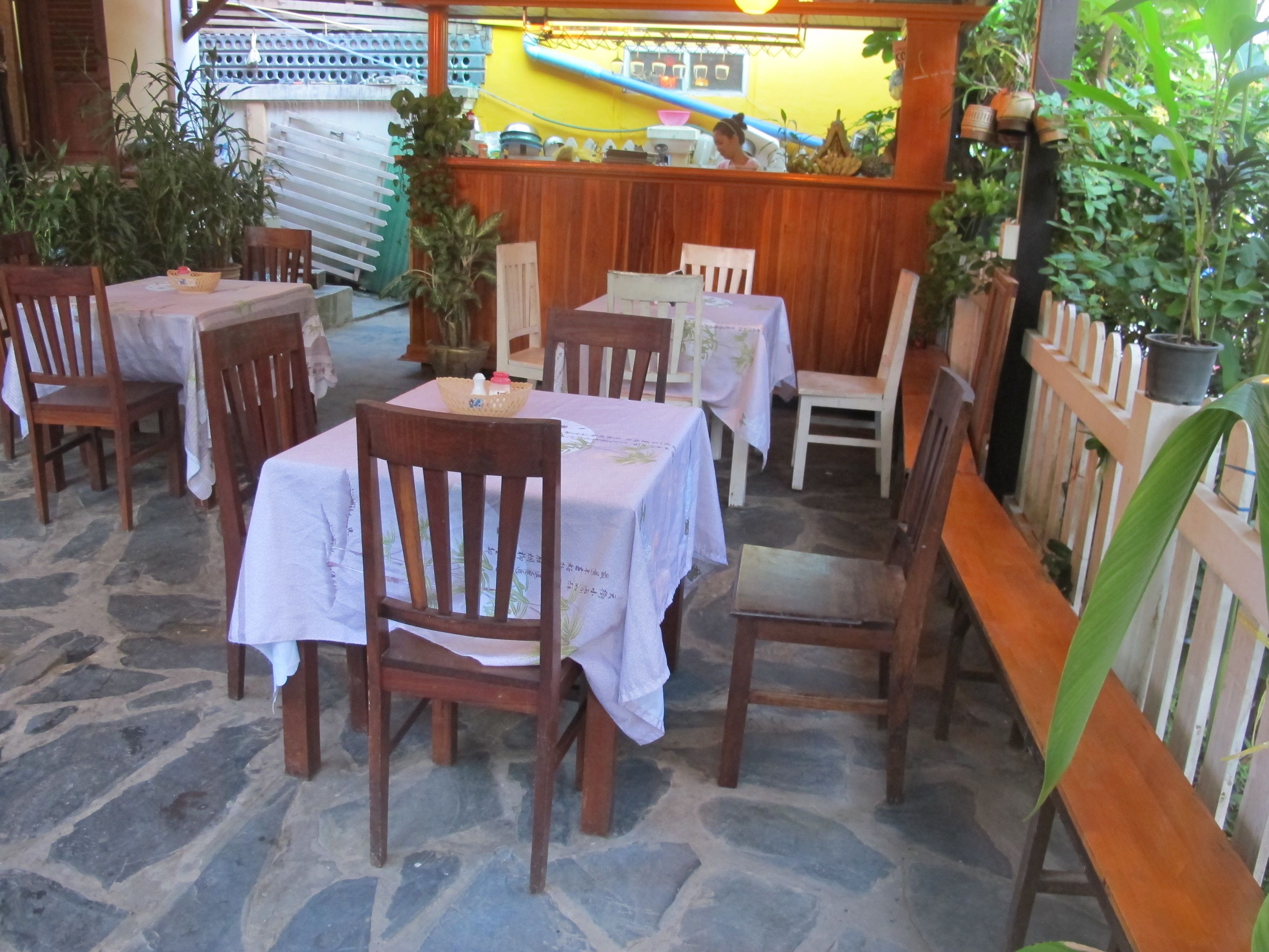 An outdoor dining area with stone flooring, wooden tables and chairs, and potted plants.